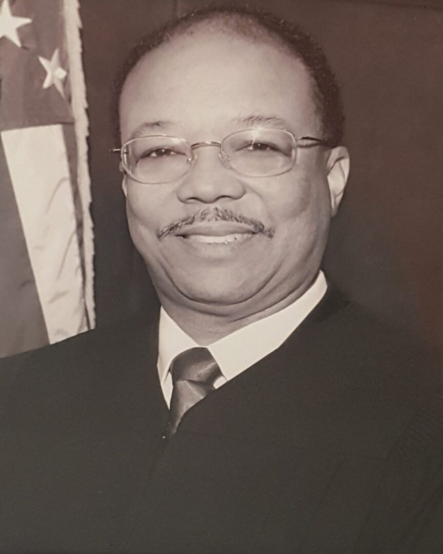 Portrait of a smiling man in formal attire with an American flag in the background.