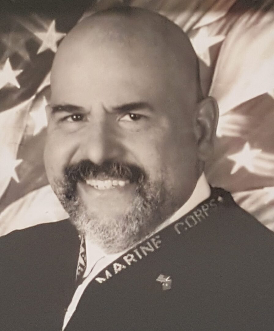 A smiling man with a bald head and beard in front of a flag.