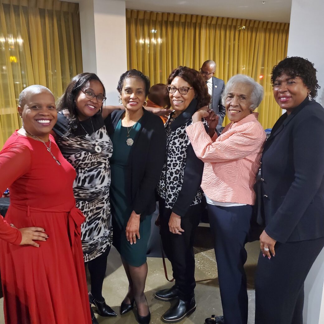 A group of six women posing together indoors, smiling warmly.
