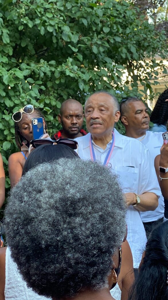 A man speaking outdoors with a group of people around him.