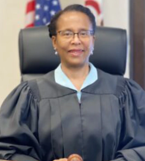 A judge in robes sitting in a courtroom with flags in the background.