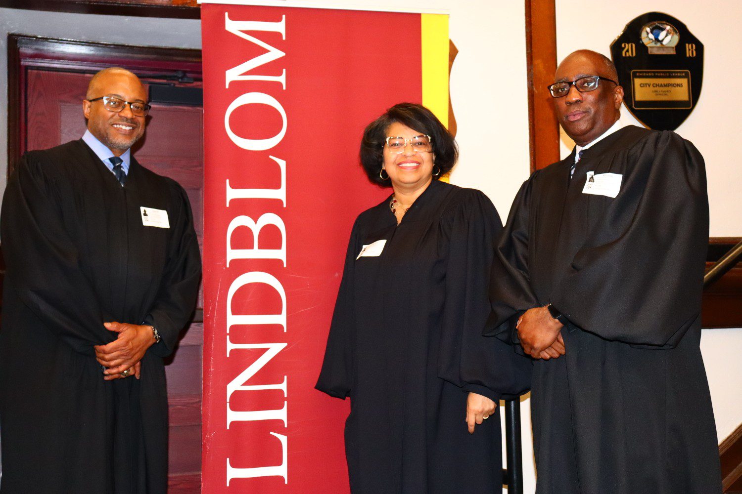 Three people in formal attire stand by a Lindblom banner indoors.