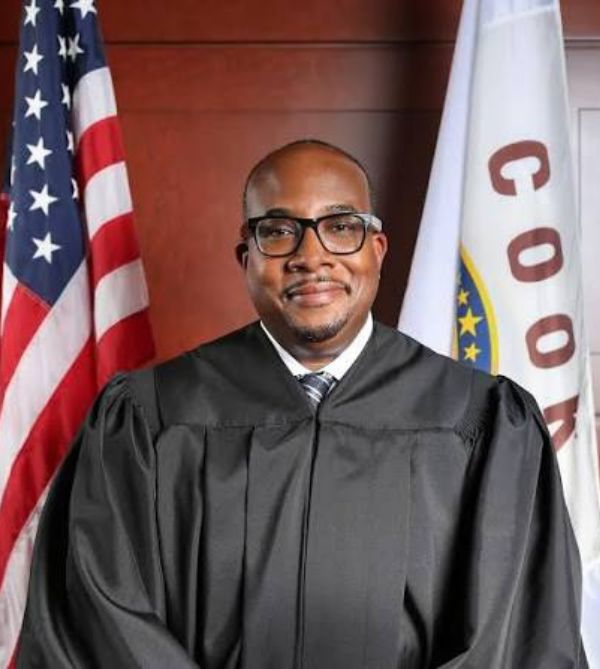 A judge in black robe standing with flags in the background.