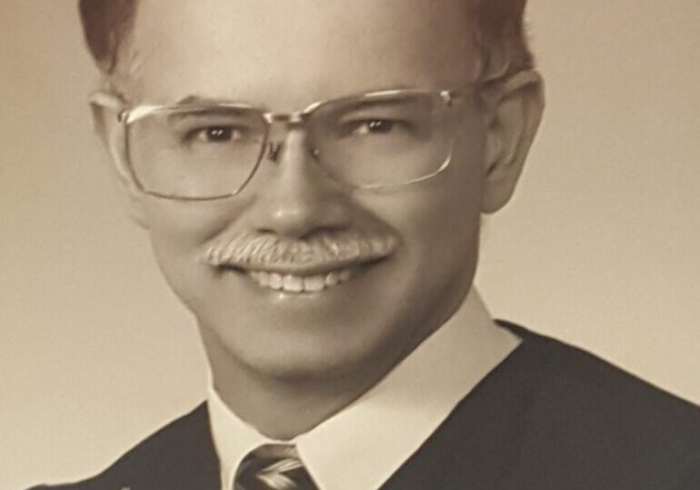 Portrait of a smiling man with glasses and a mustache in formal attire.